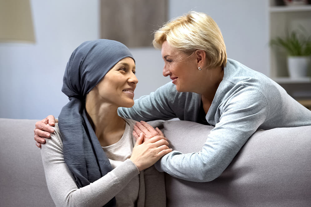 Krebspatientin und einer weitere Frau lächeln sich gegenseitig an. Die Frau hält die Krebspatientin an den Schultern. Krebspatientin trägt eine Kopfbedeckung - Krebsnachsorge bei Tumorleiden, Mannheim